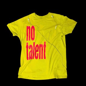May include: A lime green short-sleeved t-shirt featuring the red text "no talent" printed on the front. A pair of white wired earbuds is draped over the neckline, with the thin white cord extending down the front of the shirt.