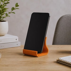 Könnte beinhalten: Ein leuchtend orangefarbener Smartphone-Ständer mit geripptem, wellenförmigem Design, der ein schwarzes Smartphone in vertikaler Position hält. Der Ständer verfügt über eine geschwungene Basis und eine gerillte Stützstruktur für das Gerät.