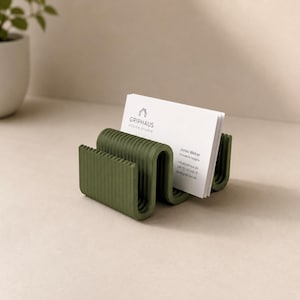 Könnte beinhalten: Ein olivgrüner, wellenförmiger Visitenkartenhalter mit gerippter Textur. Er enthält einen Stapel weißer Visitenkarten mit dem Text GRIPHAUS DESIGN STUDIO, Jonas Weber, Founder & Designer, sowie Kontaktdaten inklusive einer Telefonnummer.