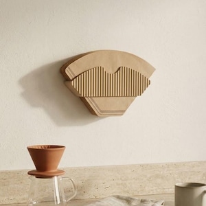 Könnte beinhalten: Ein an der Wand montierter, beigefarbener Papierkaffeefilterhalter mit gerippter Textur, platziert über einem Kaffeefilter aus Terrakotta auf einer Glaskaraffe, einem gefalteten beigen Tuch, einer Keramiktasse und einem schwarzen Schwanenhalskessel.