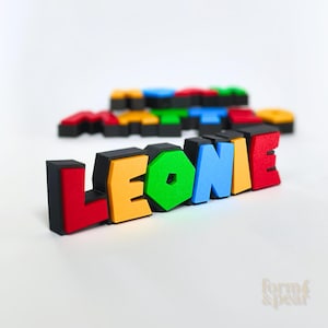 Op de afbeelding: Een kleurrijke, 3D-geprinte naam, "LEONIE", in vette blokletters. Elke letter heeft een andere kleur: rood, geel, groen, blauw en oranje, met zwarte randen. De naam staat schuin, met andere namen wazig op de achtergrond.