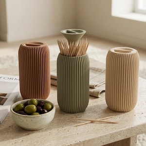 Può includere: Tre contenitori cilindrici con texture a coste verticali in terracotta, verde salvia e beige. Il contenitore verde salvia contiene stuzzicadenti in legno con un piccolo coperchio a imbuto. Una piccola ciotola maculata piena di olive verdi è vicina.