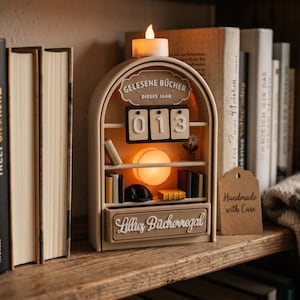 Può includere: Un bancone beige a forma di libreria ad arco con una luce sulla parte superiore, che mostra il testo Gelesene Bücher dieses Jahr e Lillies Bücherregal. Presenta un display con il numero 013, libri in miniatura e un piccolo fiore.