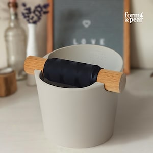 Könnte beinhalten: Ein hellgrauer Kaffeesatzbehälter mit einem horizontalen Holzstab, der eine schwarze, strukturierte Silikonhülle aufweist. Der Markenname form & pear ist auf der Vorderseite des Behälters eingeprägt. Das Design umfasst einen zylindrischen Körper.
