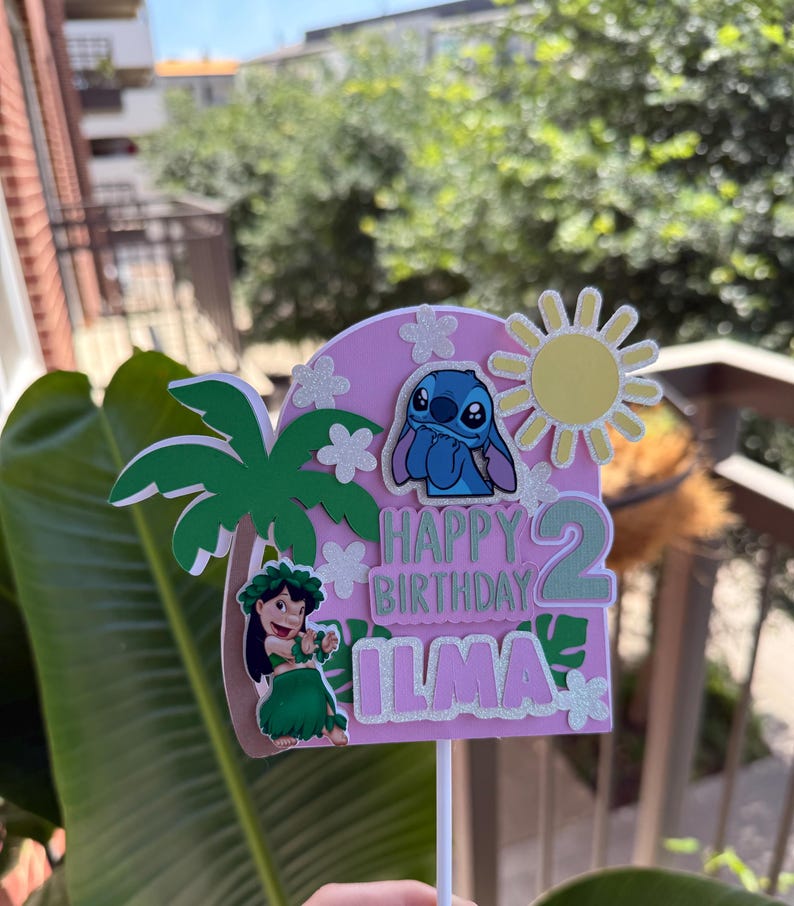 Puede incluir: Un adorno rosa para tarta de cumplea&ntilde;os con el personaje Stitch, una palmera y un sol. El texto dice "Happy Birthday Ilma 2". El adorno est&aacute; decorado con purpurina y detalles florales, ideal para una fiesta tem&aacute;tica.