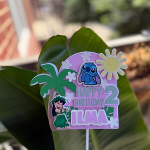 Puede incluir: Un adorno rosa para tarta de cumplea&ntilde;os con tem&aacute;tica tropical. Presenta un personaje de dibujos animados, una palmera y las palabras "Happy Birthday Ilma 2". El adorno se sujeta con un palo blanco.