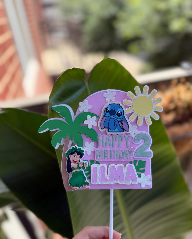 Puede incluir: Un adorno rosa para tarta de cumplea&ntilde;os con tem&aacute;tica tropical. Presenta un personaje de dibujos animados, una palmera y las palabras "Happy Birthday Ilma 2". El adorno se sujeta con un palo blanco.