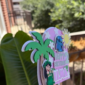 Puede incluir: Un adorno para tarta de cumplea&ntilde;os rosa y blanco con una caricatura de Stitch, una palmera y las palabras "Happy Birthday Ilma". El adorno tiene acentos de sol y flores.