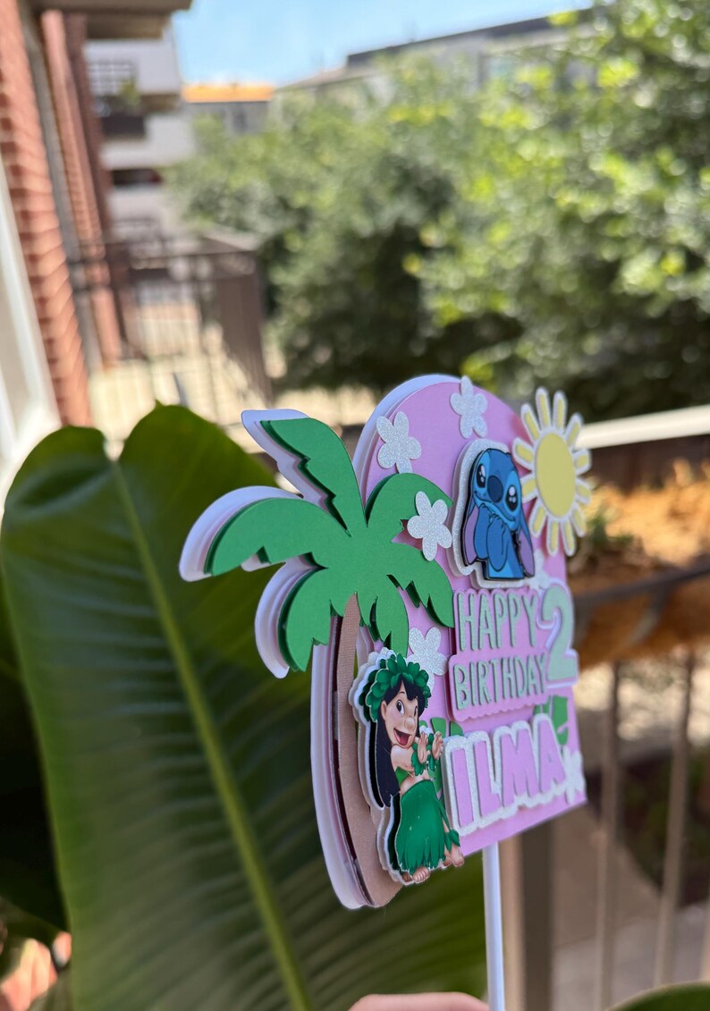 Puede incluir: Un adorno para tarta de cumplea&ntilde;os rosa y blanco con una caricatura de Stitch, una palmera y las palabras "Happy Birthday Ilma". El adorno tiene acentos de sol y flores.