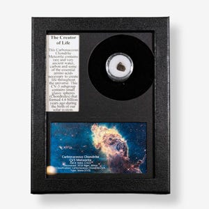 Pode incluir: Uma exibição emoldurada com um meteorito condrito carbonáceo. A moldura inclui texto descrevendo a composição e a origem do meteorito, juntamente com uma imagem colorida de uma nebulosa. O meteorito está envolto em um pequeno recipiente transparente.
