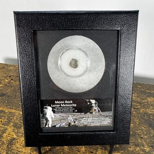 May include: A black framed display containing a lunar meteorite. The frame holds a circular, clear container with a small, grey rock. Below the rock is a printed label with text about the meteorite, including its name and origin. The frame is on a wooden surface.