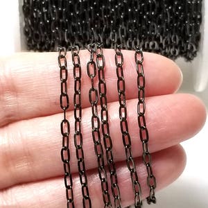 May include: Black chain in a spool, with several strands of chain laid out on a hand. The chain is a fine, delicate chain with a square link design.