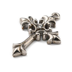 May include: Silver-toned cross pendant with ornate floral detailing and a small, clear gemstone at the center. The cross has a loop at the top for hanging and is set against a white background.