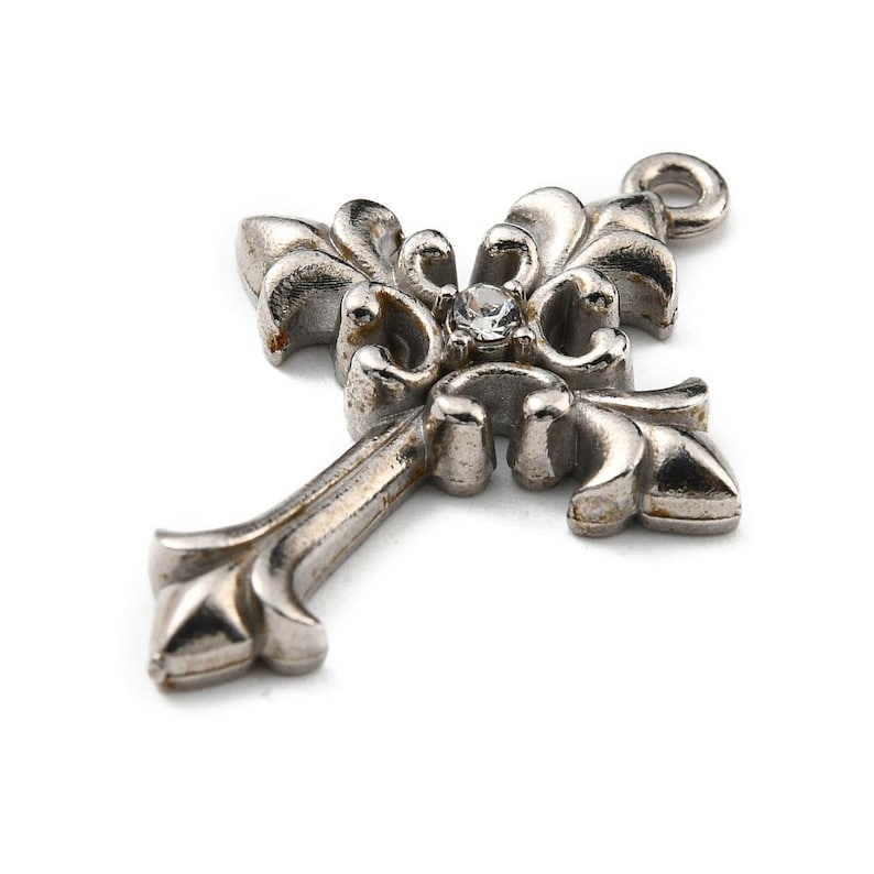 May include: Silver-toned cross pendant with ornate floral detailing and a small, clear gemstone at the center. The cross has a loop at the top for hanging and is set against a white background.