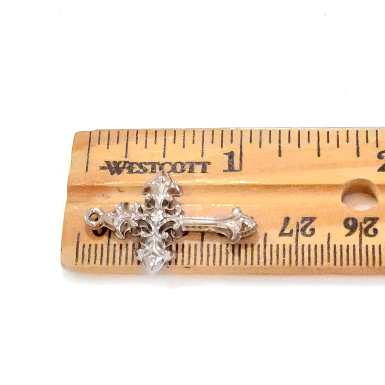 May include: A silver-colored cross with ornate floral detailing, resting on a wooden ruler. The cross is approximately 1 inch in length, showcasing intricate design elements. The ruler's markings are clearly visible, providing a size reference.