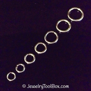 May include: A set of eight silver jump rings of varying sizes, arranged in a diagonal line on a black background.  The rings are all smooth and shiny, and they are perfect for jewelry making.