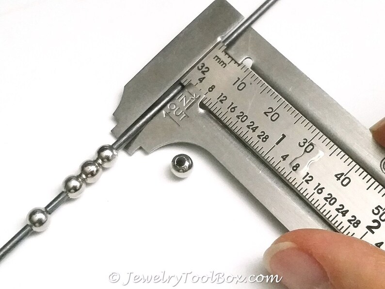May include: A close-up of a metal ruler measuring a silver bead. The ruler is marked in millimeters and inches. The bead is about 4 millimeters in diameter.