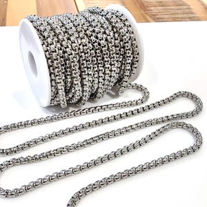 May include: A spool of shiny, silver-colored chain. The chain is a round, box-style design, with a portion unspooled and lying flat. The chain is likely made of stainless steel or a similar metal. The background is white.
