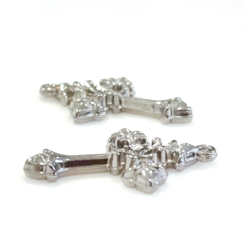 May include: Two silver-colored cross pendants with ornate floral designs. The crosses have a detailed, textured appearance and a small loop at the top for attaching to a chain or other jewelry component. The crosses are displayed on a white background.