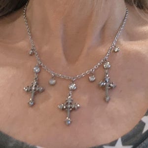 May include: Silver-toned necklace featuring three ornate cross pendants and several small, sparkling accents. The necklace is worn around the neck and is visible against a neutral background.