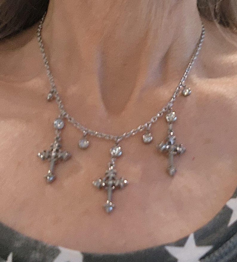 May include: Silver-toned necklace featuring three ornate cross pendants and several small, sparkling accents. The necklace is worn around the neck and is visible against a neutral background.