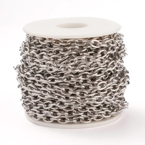 May include: A spool of silver-colored chain, wound tightly around a white plastic spool. The chain is made of metal links, and the spool is sitting on a white surface.