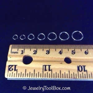 May include: A set of seven silver jump rings of varying sizes, measured in millimeters on a wooden ruler. The jump rings are arranged in order of size, with the smallest ring on the left and the largest ring on the right.