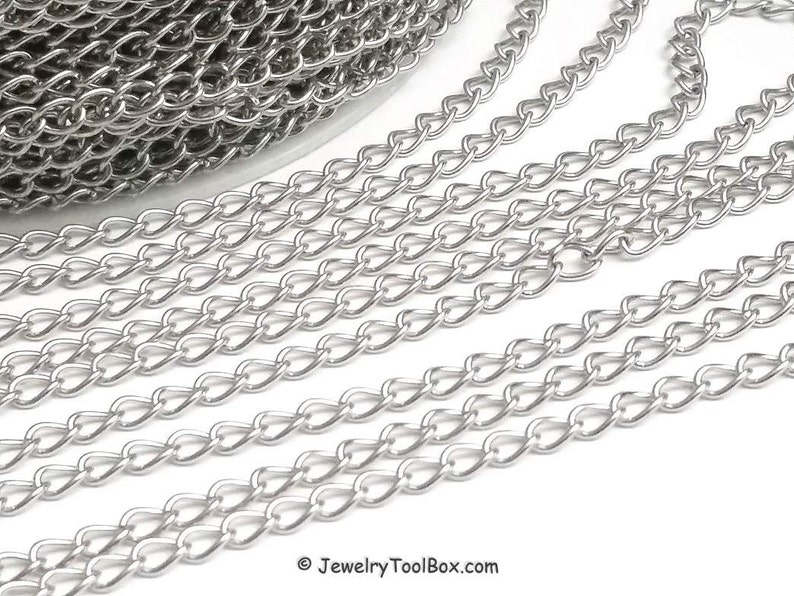 May include: Silver curb chain, a type of chain with flat, oval links, is shown in a close-up image. The chain is laid out in a straight line, with multiple strands overlapping.