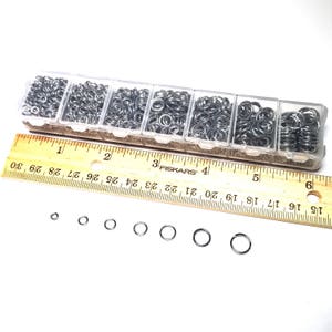 May include: A clear plastic container with six compartments filled with silver jump rings of various sizes. A wooden ruler with the text "FISKARS" is below the container.  The ruler is marked in inches.