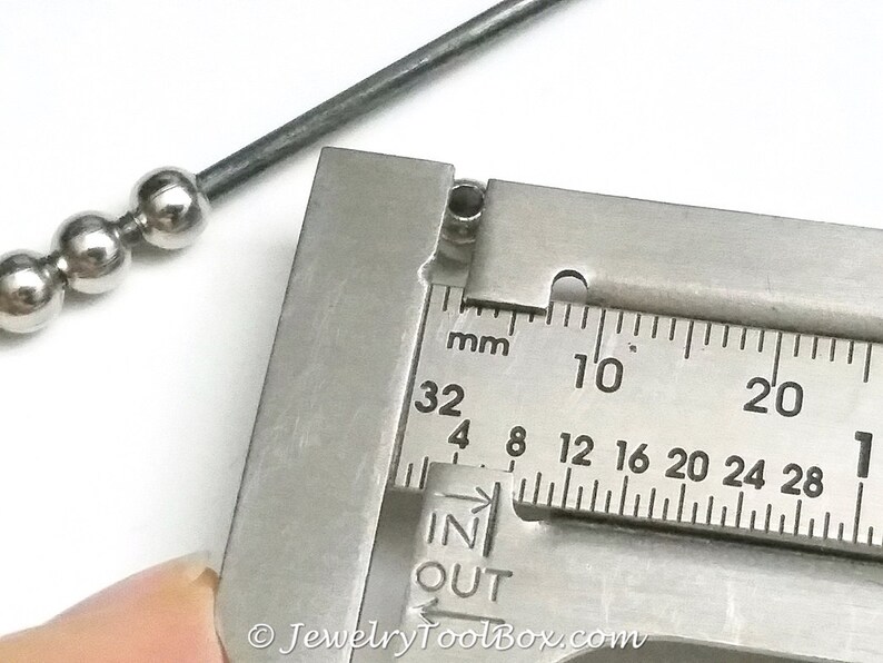 May include: Close-up of a silver metal bead measured with a caliper. The caliper reads 32 mm, 4 mm, 8 mm, 12 mm, 16 mm, 20 mm, 24 mm, 28 mm, 10, 20, and 1. The caliper has the words "IN" and "OUT" printed on it.