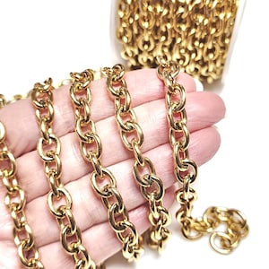 May include: Gold chain link with a smooth, oval-shaped design. The chain is shown in a hand, with a spool of the chain in the background.