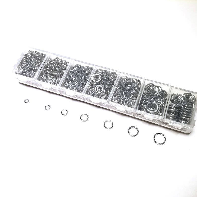 May include: A clear plastic container with six compartments filled with silver jump rings of various sizes. The jump rings are arranged in order of size, with the smallest rings in the first compartment and the largest rings in the last compartment.