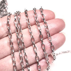 May include: A close-up of five silver chains of different lengths, with a hand holding the chains in place. The chains are made of small, oval-shaped links.
