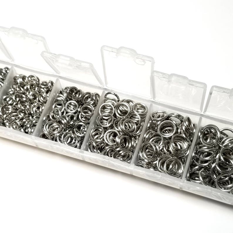 May include: A clear plastic storage box filled with six compartments, each containing a large quantity of silver jump rings.