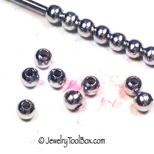 May include: A close-up of a group of silver metal beads. The beads are round and have a smooth surface. There is a tool with a row of beads in the background.