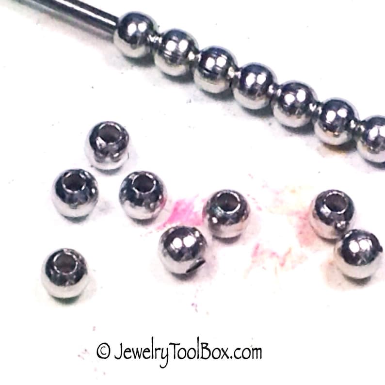 May include: A close-up of a group of silver metal beads. The beads are round and have a smooth surface. There is a tool with a row of beads in the background.