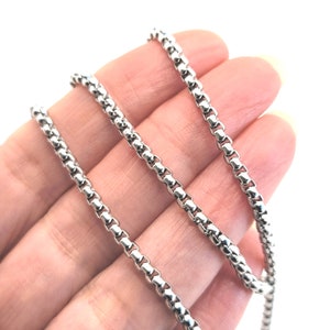 May include: A silver chain necklace with a box chain design. The chain is shown in three layers on a hand.