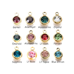 May include: Assortment of 12 gold-tone charms with different colored gemstones. The charms are round and have a small loop at the top for attaching to a necklace or bracelet. The gemstones include garnet, amethyst, aquamarine, crystal, emerald, alexandrite, light siam, peridot, sapphire, light rose, light topaz, and blue zircon.