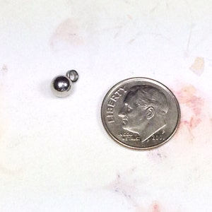 Puede incluir: Un abalorio de metal plateado en forma de bola con un bucle para sujetarlo a un collar o pulsera. El abalorio es aproximadamente del tama&ntilde;o de una moneda de diez centavos.