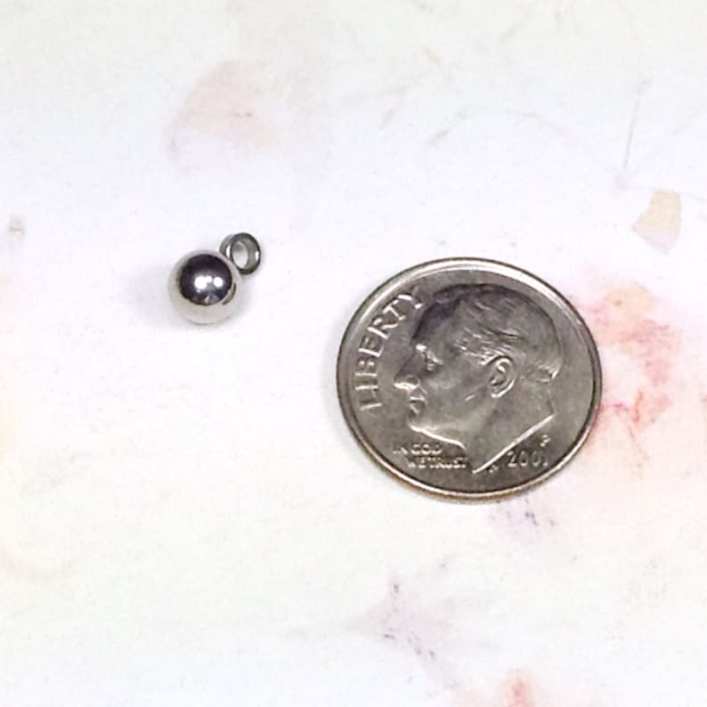 Puede incluir: Un abalorio de metal plateado en forma de bola con un bucle para sujetarlo a un collar o pulsera. El abalorio es aproximadamente del tama&ntilde;o de una moneda de diez centavos.