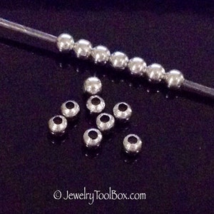 May include: A close-up of silver metal beads. The beads are round and have a smooth finish. There are two rows of beads, one row is on a wire and the other row is scattered on a black surface.