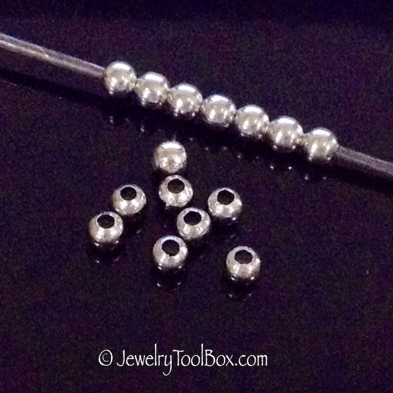 May include: A close-up of silver metal beads. The beads are round and have a smooth finish. There are two rows of beads, one row is on a wire and the other row is scattered on a black surface.