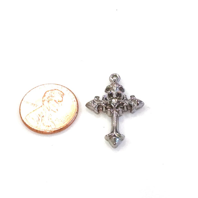 May include: A silver-colored cross pendant with ornate detailing, featuring a fleur-de-lis design at the top and a small gem in the center. The cross is next to a penny for size comparison.