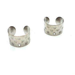 May include: Two silver ear cuffs with a star pattern. The cuffs are open and have a small hole in the center.