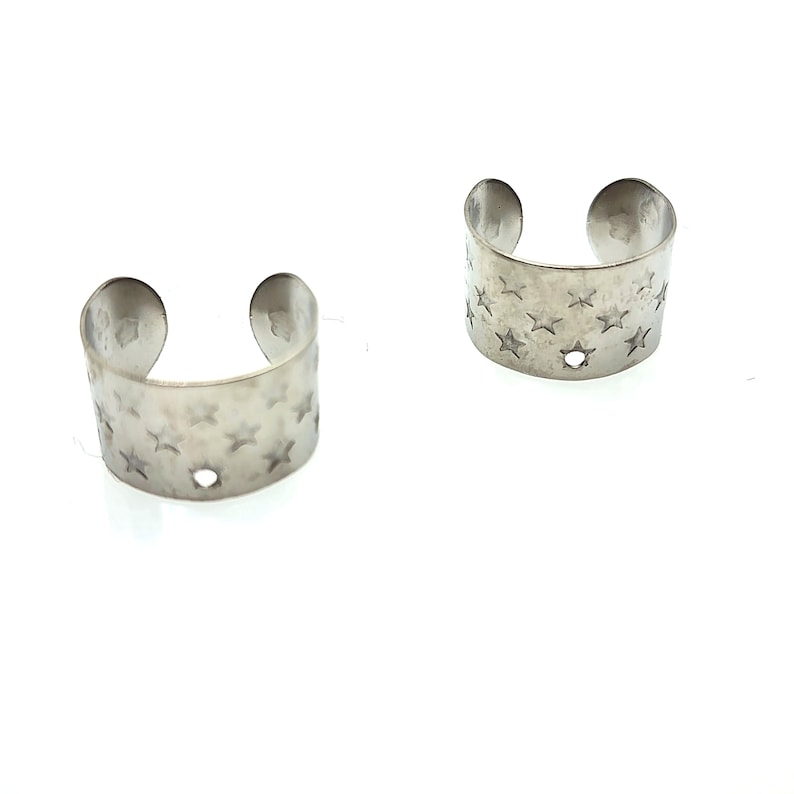 May include: Two silver ear cuffs with a star pattern. The cuffs are open and have a small hole in the center.