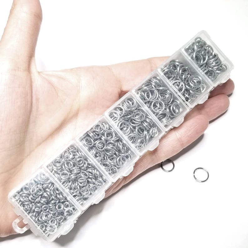 May include: A clear plastic storage box filled with six compartments containing silver jump rings. The box is held in a hand.
