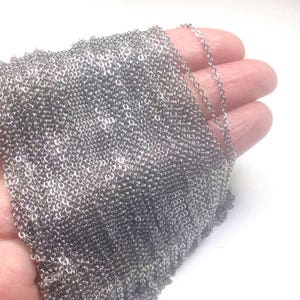 May include: A silver chain necklace, shown in a close-up image, is folded in a hand. The chain is made of small, linked metal rings.