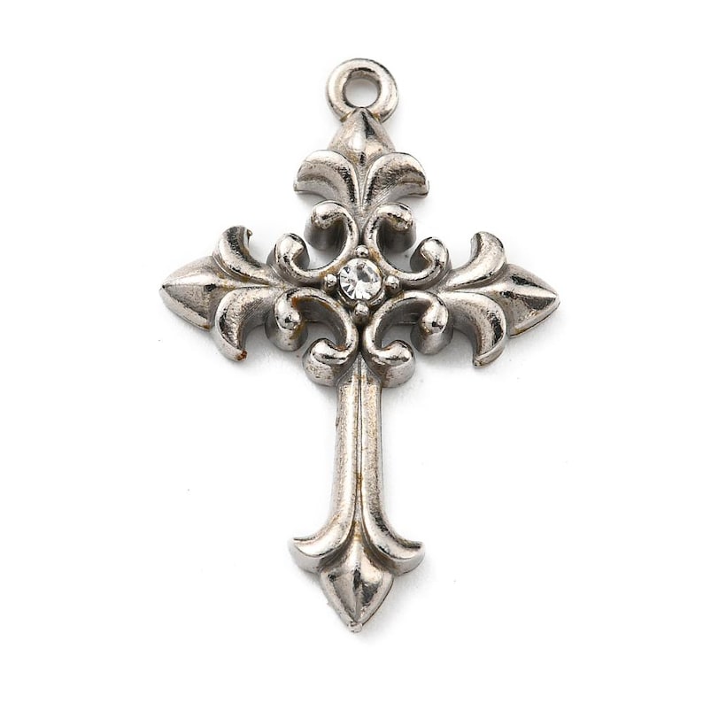 May include: Silver-toned metal cross pendant with ornate fleur-de-lis style arms and a small, clear crystal at the center. The pendant has a loop at the top for attaching to a chain or cord. The cross is a religious symbol.