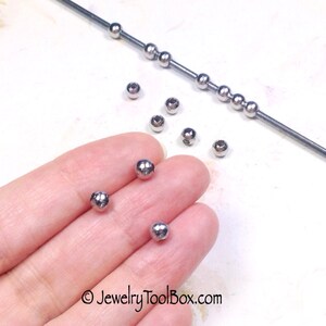 May include: A close-up of silver metal beads, some are on a wire, others are on a hand. The beads are round and smooth.