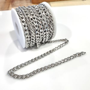 May include: A roll of silver-colored curb chain sits next to a length of the same chain. The chain is made of small, interlocking links. The roll is on a white spool. The background is white and light brown.